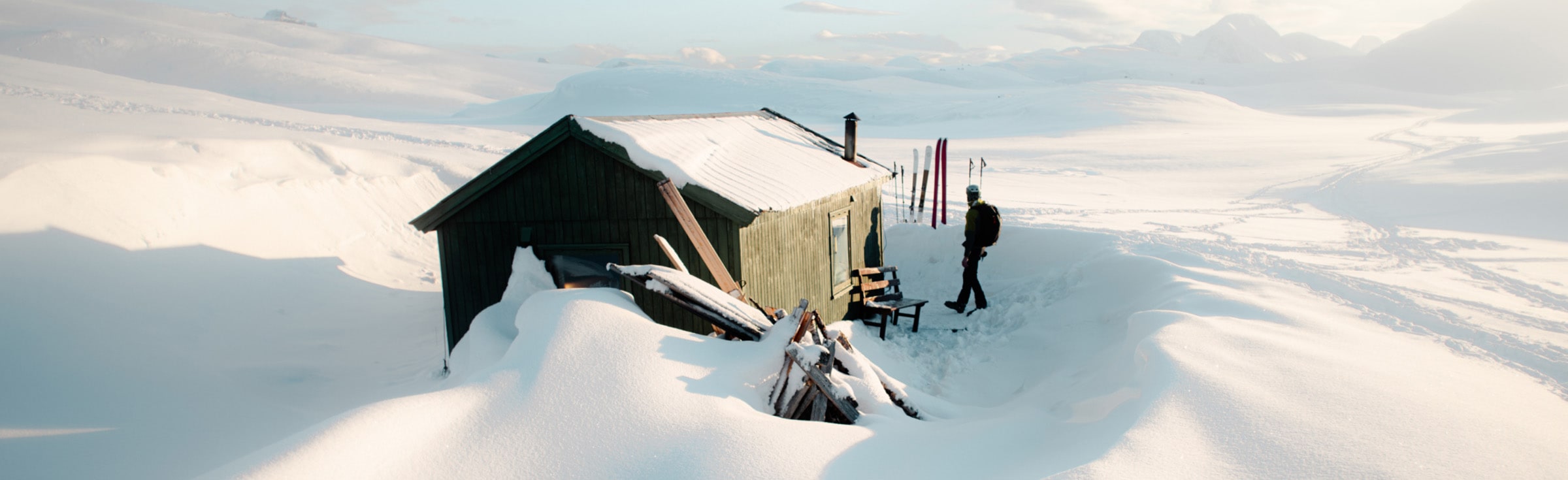 a chalet partially covered in snow, skis planted in front and a guy walking in