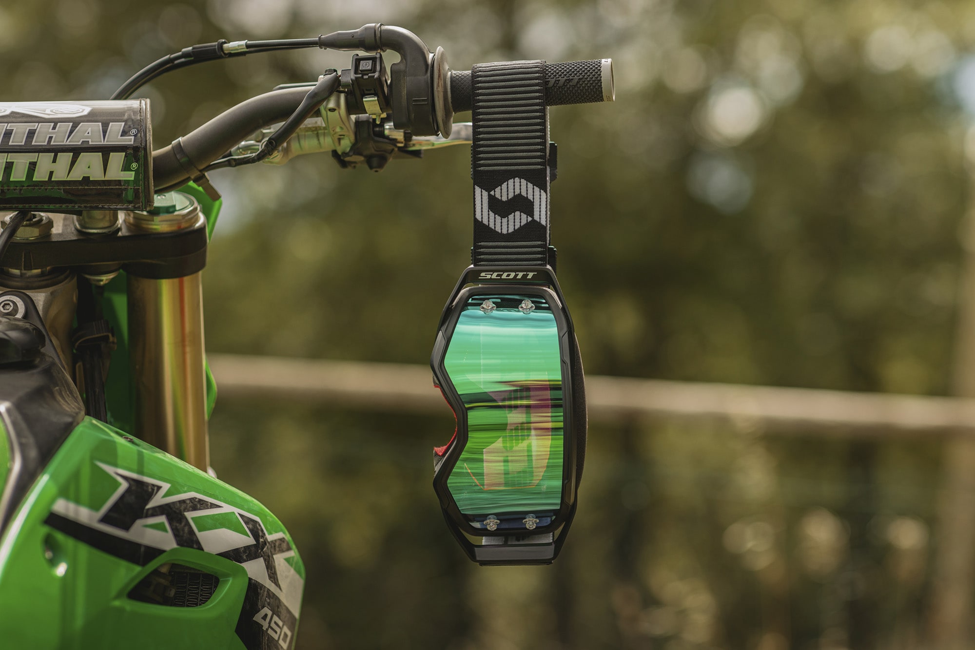 black goggle with a green chrome screen hanging to a green motorbike handlebar