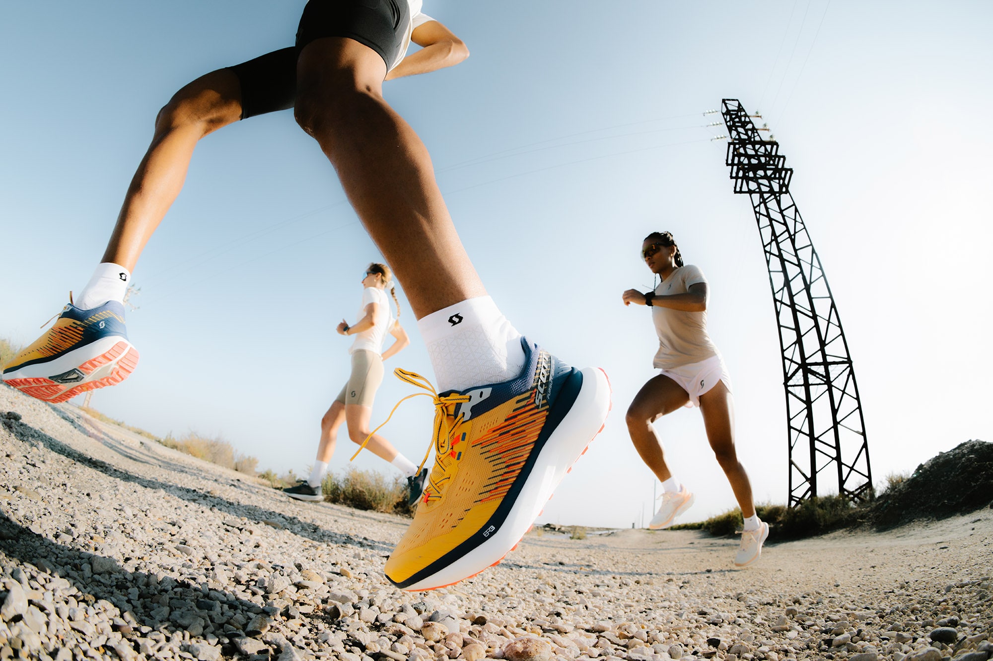 Three runners wearing SCOTT Sports apparel and Pursuit Gravel shoes in mid-stride on a gravel path, capturing energy, performance, and trail running experience.
